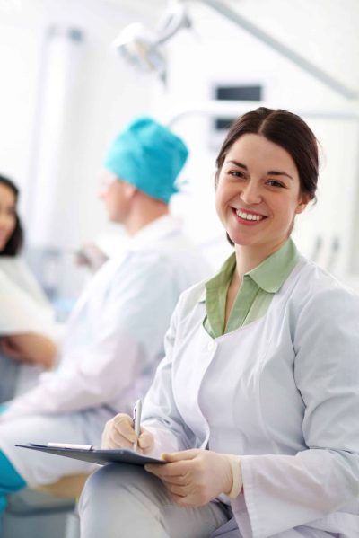 Smiling nurse with clipboard looking at camera on background of her colleague